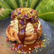 A cream puff covered in melted chocolate on a black earthen plate and sprikled with nuts and colorful accoutrements courtesy of Hello Stranger Manila