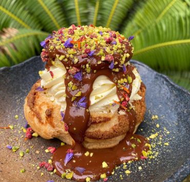 A cream puff covered in melted chocolate on a black earthen plate and sprikled with nuts and colorful accoutrements courtesy of Hello Stranger Manila
