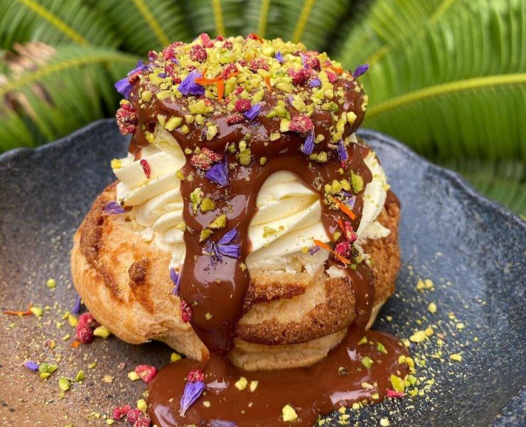 A cream puff covered in melted chocolate on a black earthen plate and sprikled with nuts and colorful accoutrements courtesy of Hello Stranger Manila